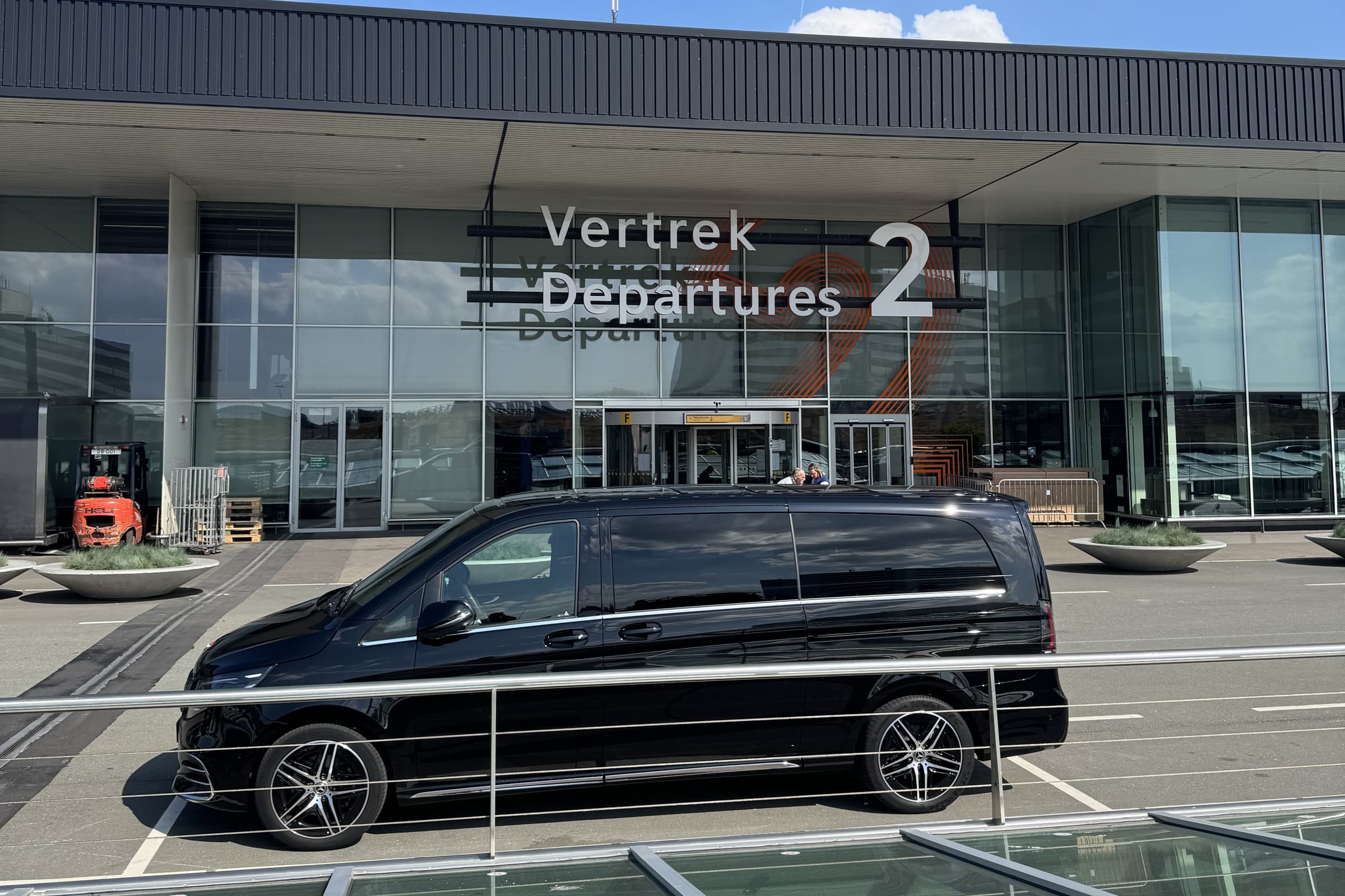 Zwarte Mercedes V-Class van Taxi Centrale Heemskerk voor de glazen gevel van Vertrekhal 2 op Schiphol, onder het bord Vertrek Departures 2